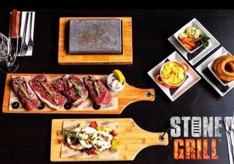 A volcanic hot stone on its wooden board, laid out with sliced steaks, sides and the Stone Grill mark on a dark table