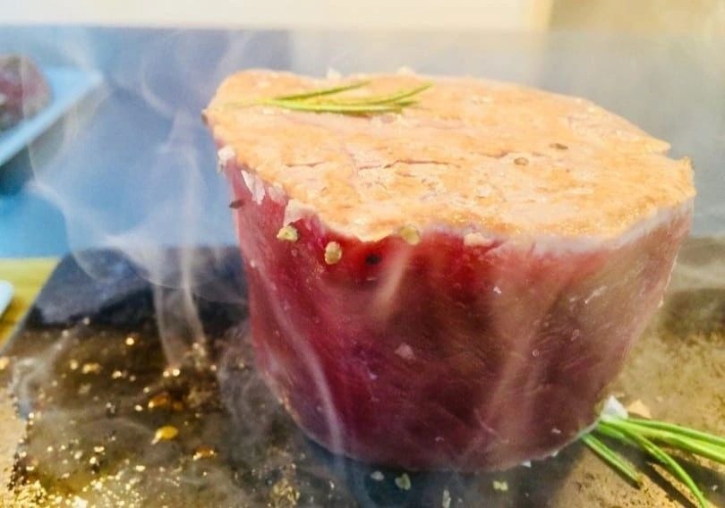 A thick cut of meat with rosemary and coarse salt sizzling on the hot stone, smoke rising