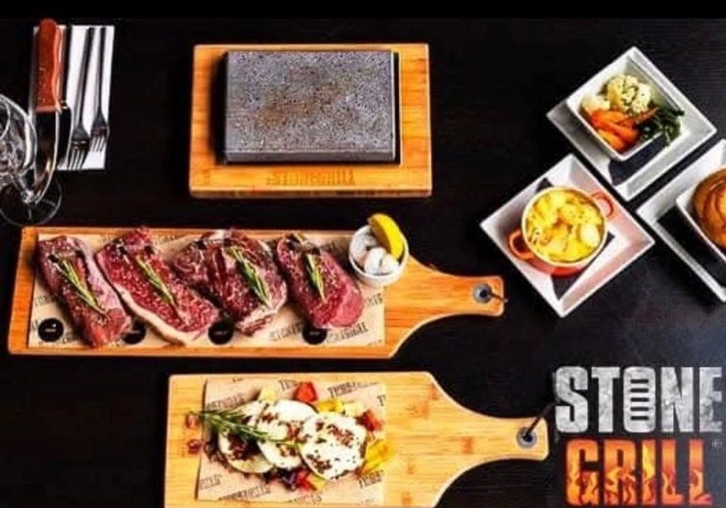 A volcanic hot stone on its wooden board, laid out with sliced steaks, sides and the Stone Grill mark on a dark table